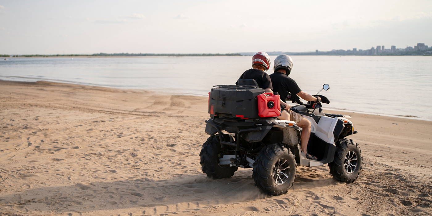 Two riders enjoy a thrilling adventure on an ATV, navigating the sandy beach along a serene coastline. The tranquil water reflects the soft afternoon light